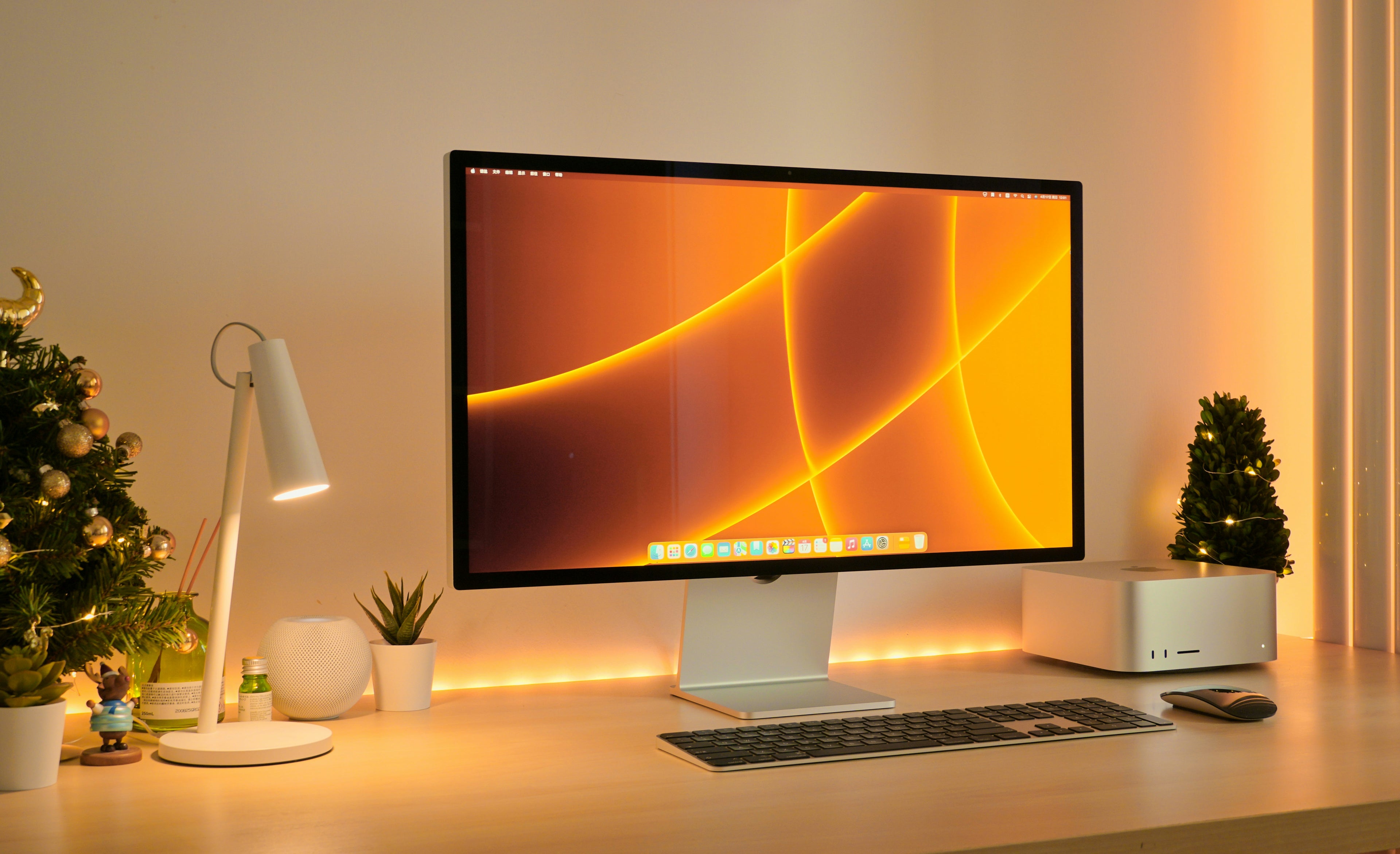 A large widescreen iMac with a silver stand sits on a wooden desk in a warmly lit home office setup, alongside a white desk lamp, small potted plants, a white computer case, and a wireless mouse.