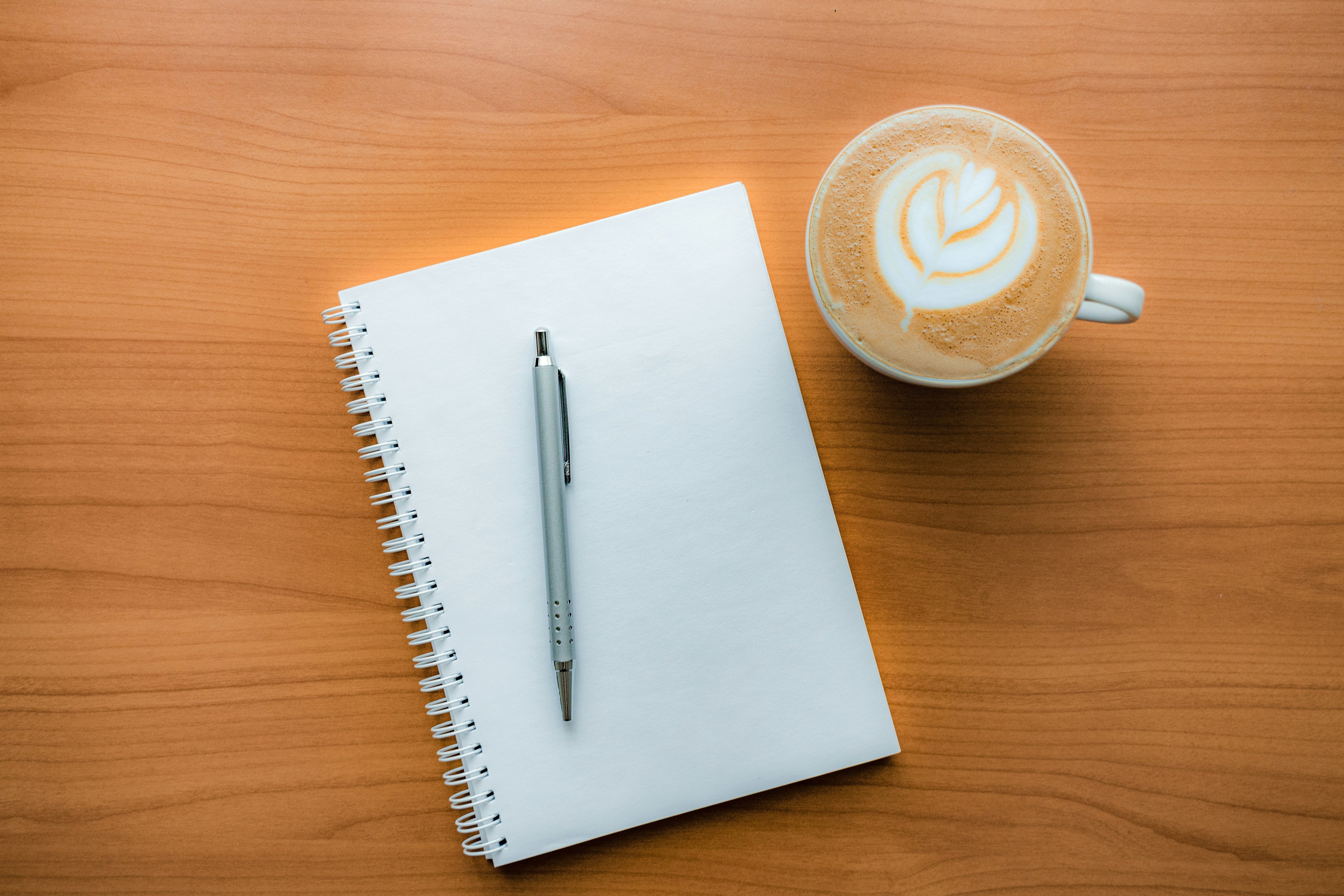 A white spiral-bound notebook with a silver pen on a warm wooden table, next to a cup of latte with leaf latte art.