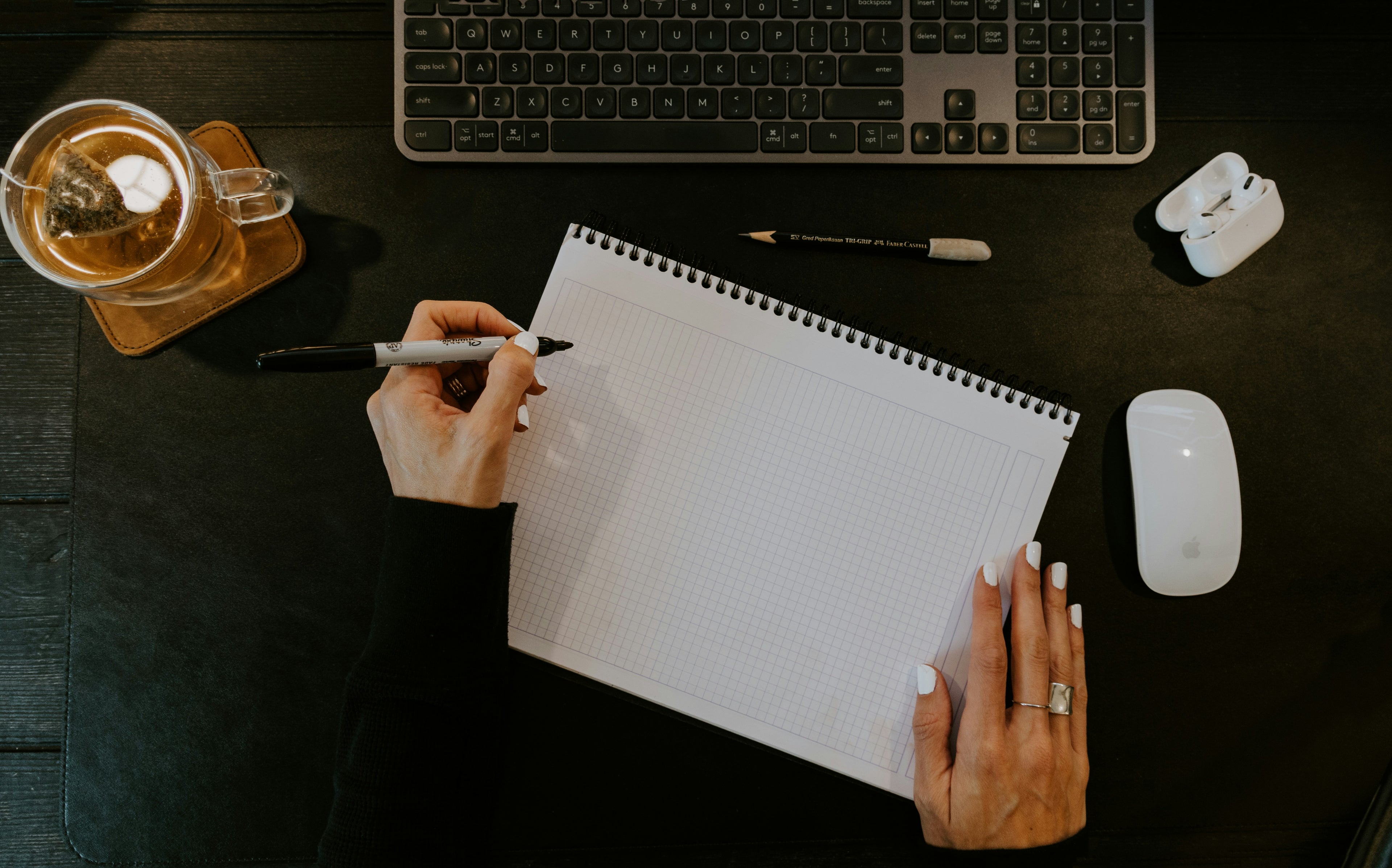 Person writing in a notebook on a desk with a keyboard, mouse, and tea.