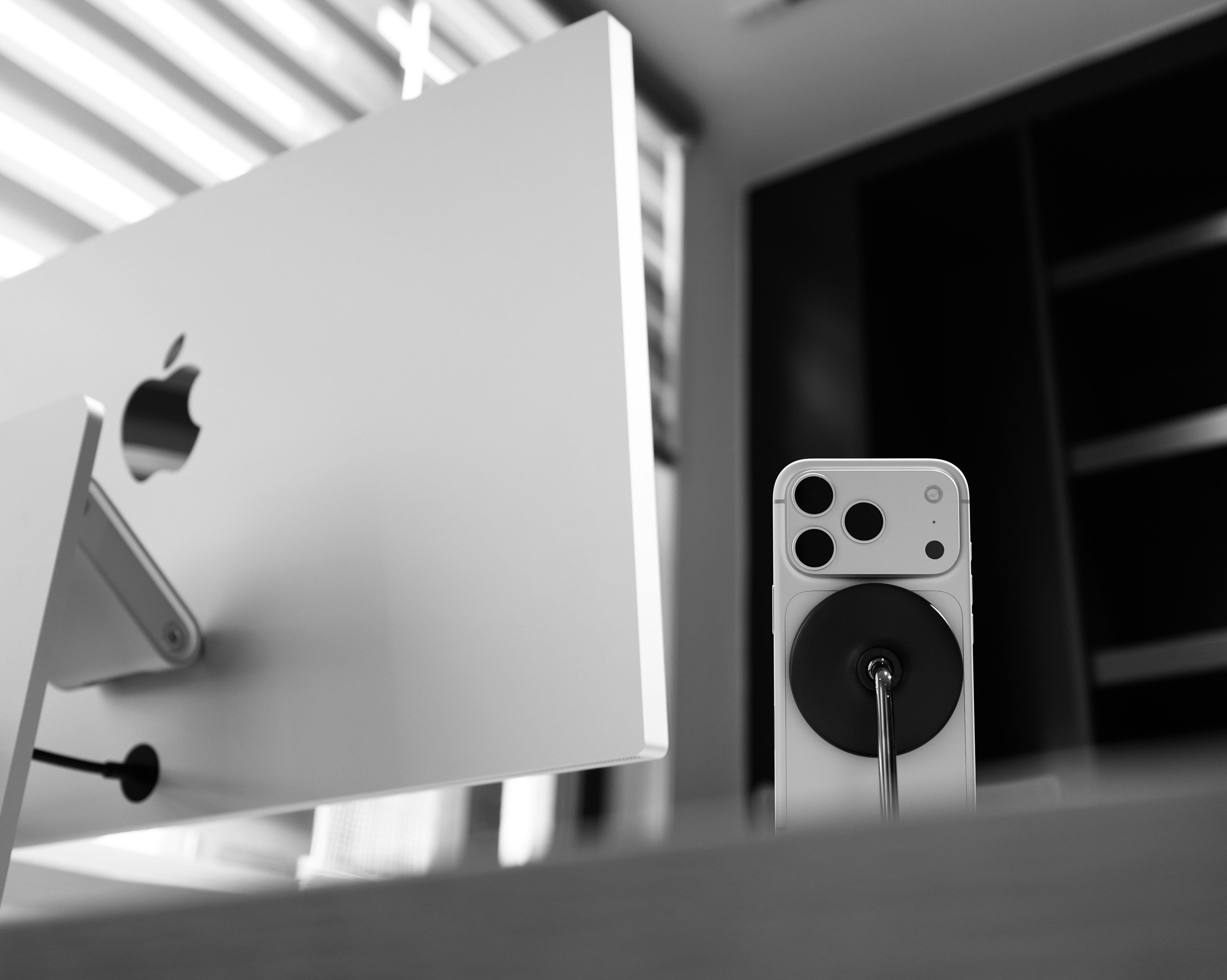 A white smartphone with a circular black magnetic accessory attached to the back, standing on a desk next to a large computer monitor in a modern office setting.